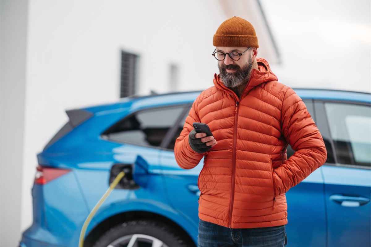 Man in an orange jacket checking his phone beside a blue electric car plugged in for charging.