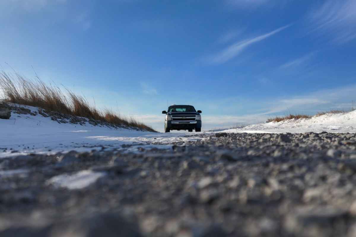 Low-angle view of an SUV driving on a snowy gravel road under a bright blue sky in Iceland.