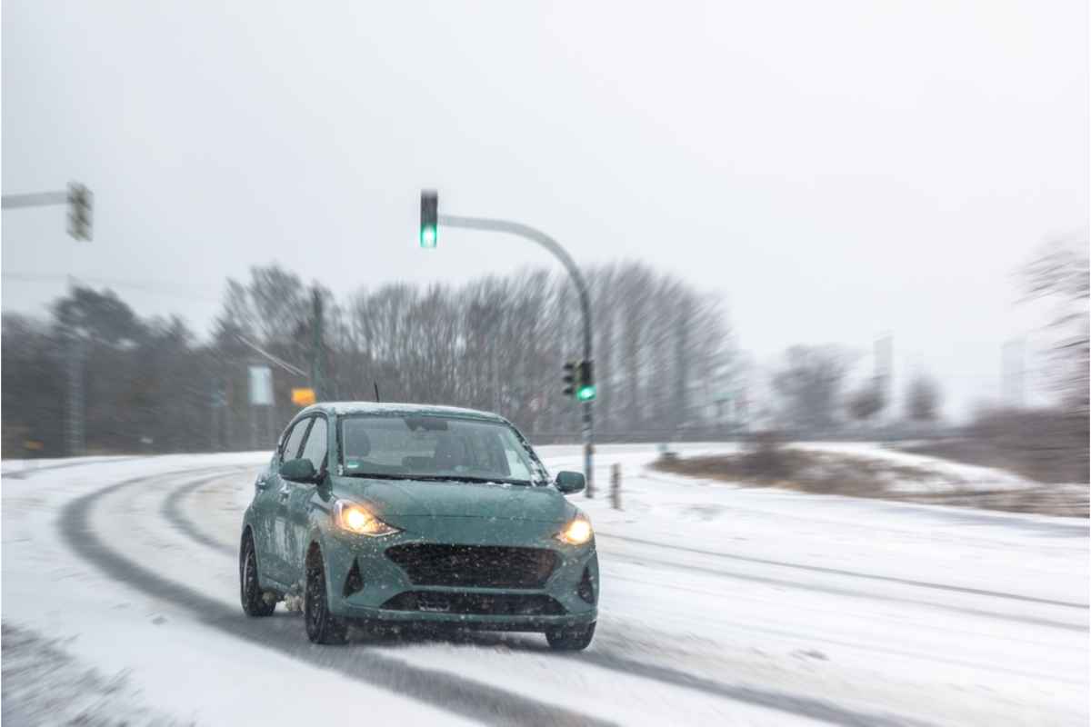 Small car driving on a snow-covered road with headlights on during winter conditions.