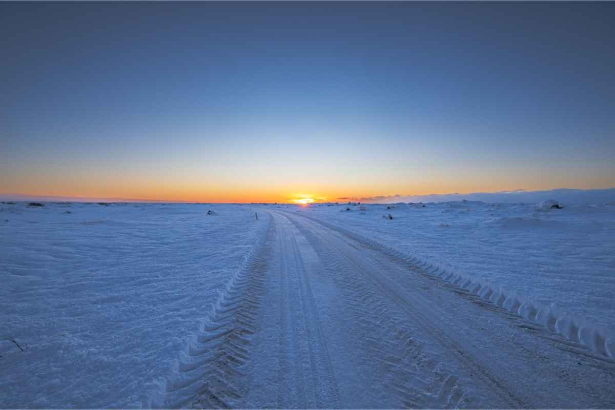 Snowy Icelandic road stretching toward the horizon at sunset under a clear sky.