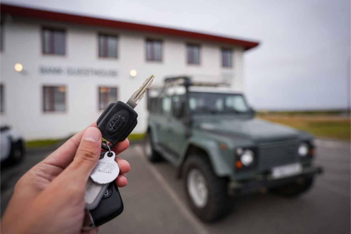 Hand holding rental car keys in front of a parked off-road vehicle outside a guesthouse.