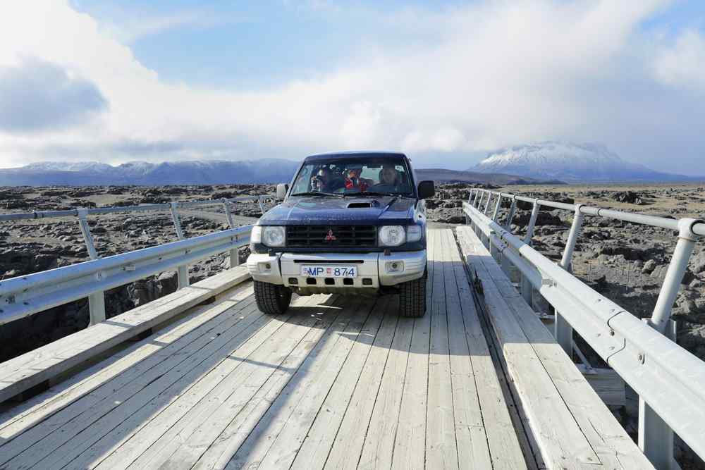 Ground clearance 4x4 vehicle crossing a narrow wooden bridge in a remote Icelandic landscape.