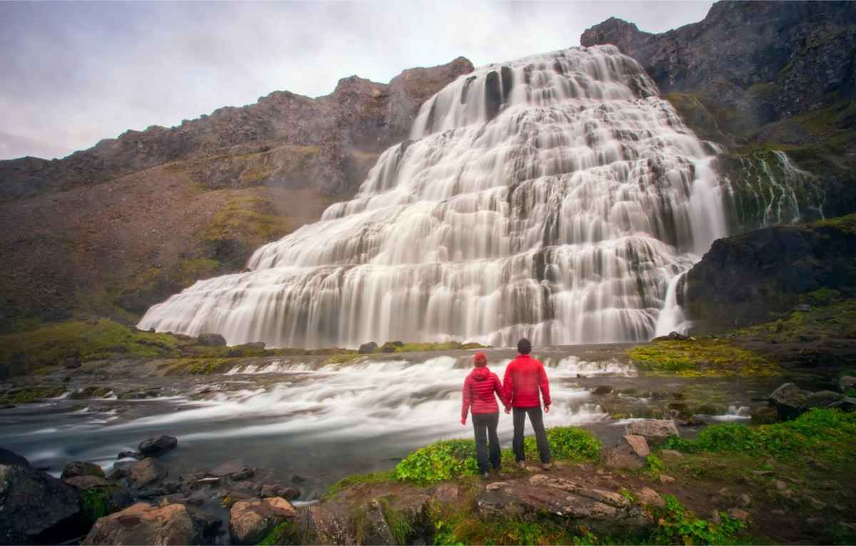 Couple in red jackets standing right in front of Dyjandi waterfall in Iceland
