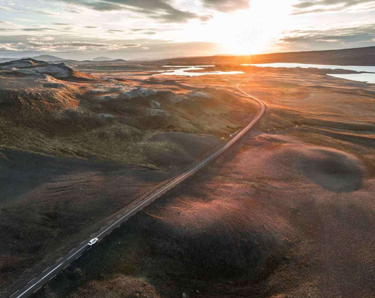 Aerial view of a car driving along a remote Iceland road at sunset, with wide volcanic landscapes and lakes in the distance.