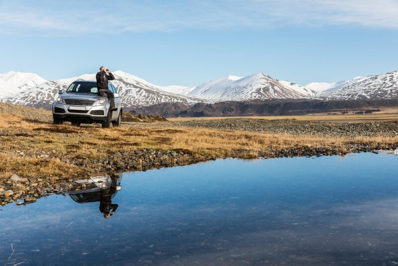 Reisender lehnt an einem weißen SUV neben einem ruhigen See in Island, in dessen Wasser sich schneebedeckte Berge spiegeln.