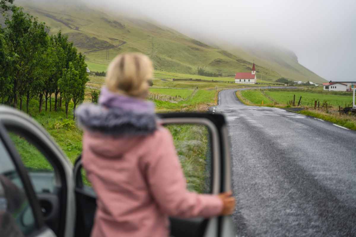 Frau steht neben ihrem Auto und betrachtet aus der Ferne eine hübsche Kapelle mit rotem Dach