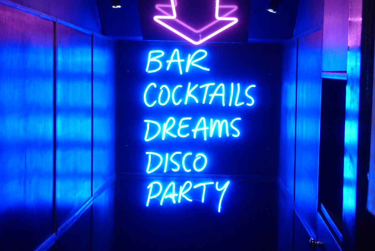 Neonschild im Flur eines Nachtclubs mit den Worten „BAR, COCKTAILS, DREAMS, DISCO, PARTY“, das in hellem Blau unter einem violetten Neonpfeil leuchtet.