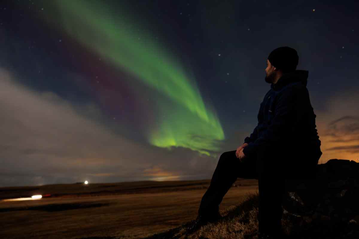 Ein Mann im Schatten genießt das wunderschöne Schauspiel der Nordlichter in Island, während er in der Nacht sitzt und warme Kleidung trägt.