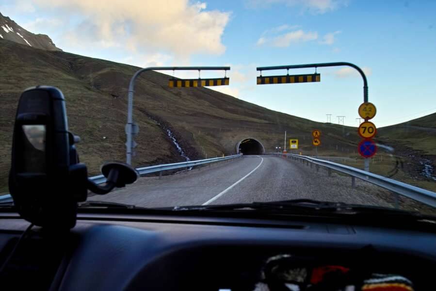 Carretera que se acerca a un túnel en Islandia vista desde el interior de un vehículo, con señales de tráfico y laderas montañosas al fondo.