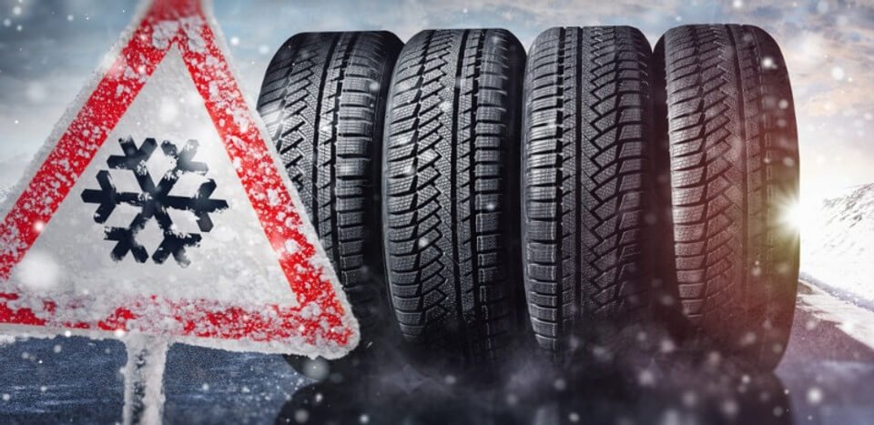 Winter road warning sign with a snowflake symbol next to four winter tires in a snowy, icy background.
