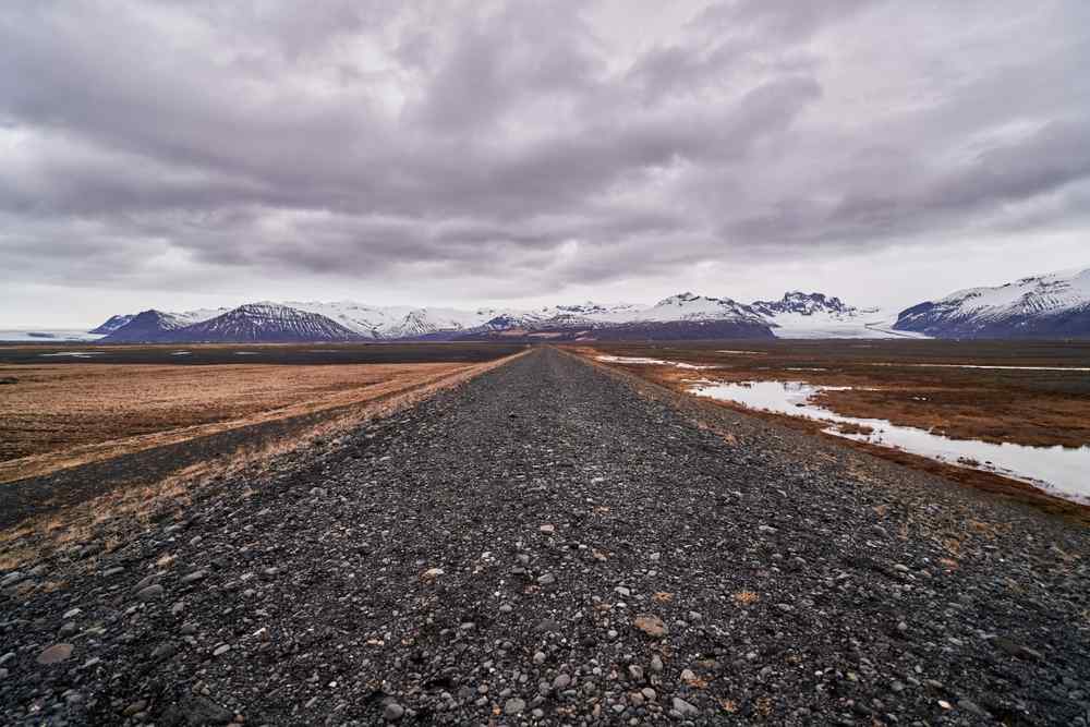 Carretera de grava vacía Carretera de grava vacía que se extiende hacia montañas nevadas bajo cielos nublados en Islandia.