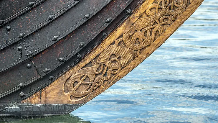 Detalle de la proa de un barco vikingo con intrincados grabados de dragones, reflejado en el agua tranquila