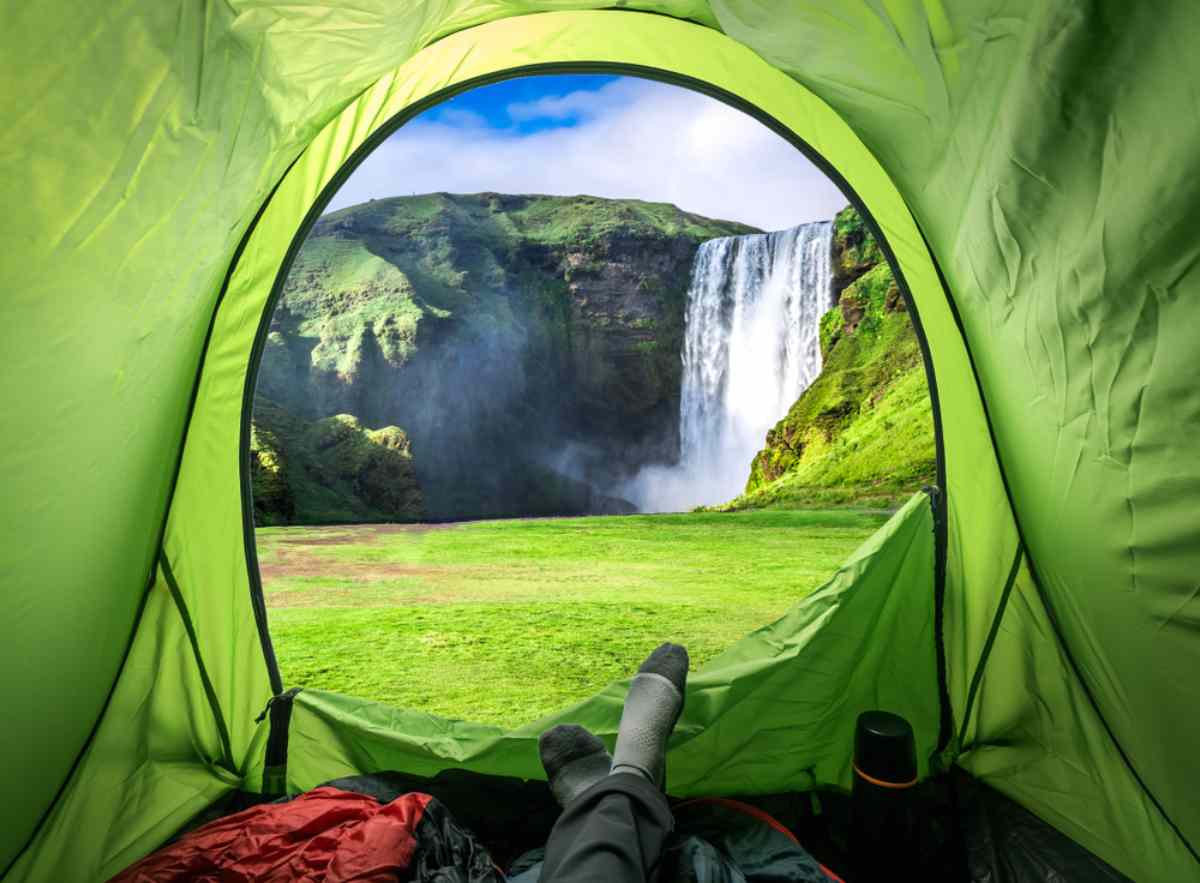 Vista desde el interior de una carpa verde, que muestra los pies de una persona en calcetines, con vistas a una impresionante cascada que cae por una exuberante montaña verde bajo un cielo azul brillante.