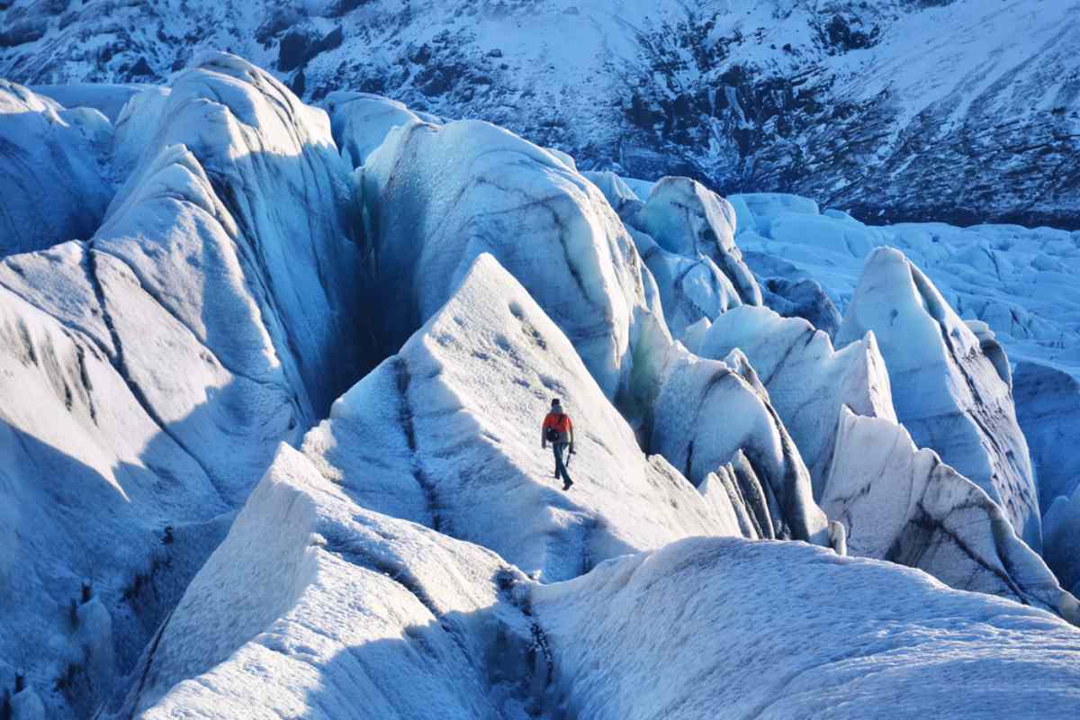 Un montañista solitario con una chaqueta roja caminando por los escarpados y helados picos de un glaciar, rodeado de vastas formaciones de hielo azul y blanco bajo un cielo despejado.