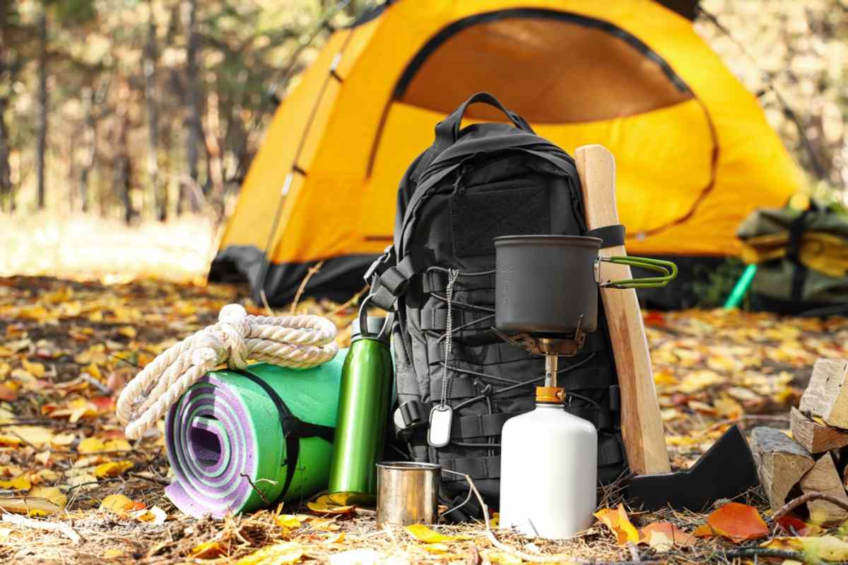 Primer plano de equipo para acampar, que incluye una mochila, un hornillo para cocinar, una colchoneta y una cuerda, sobre hojas de otoño con una tienda de campaña de color amarillo brillante de fondo.