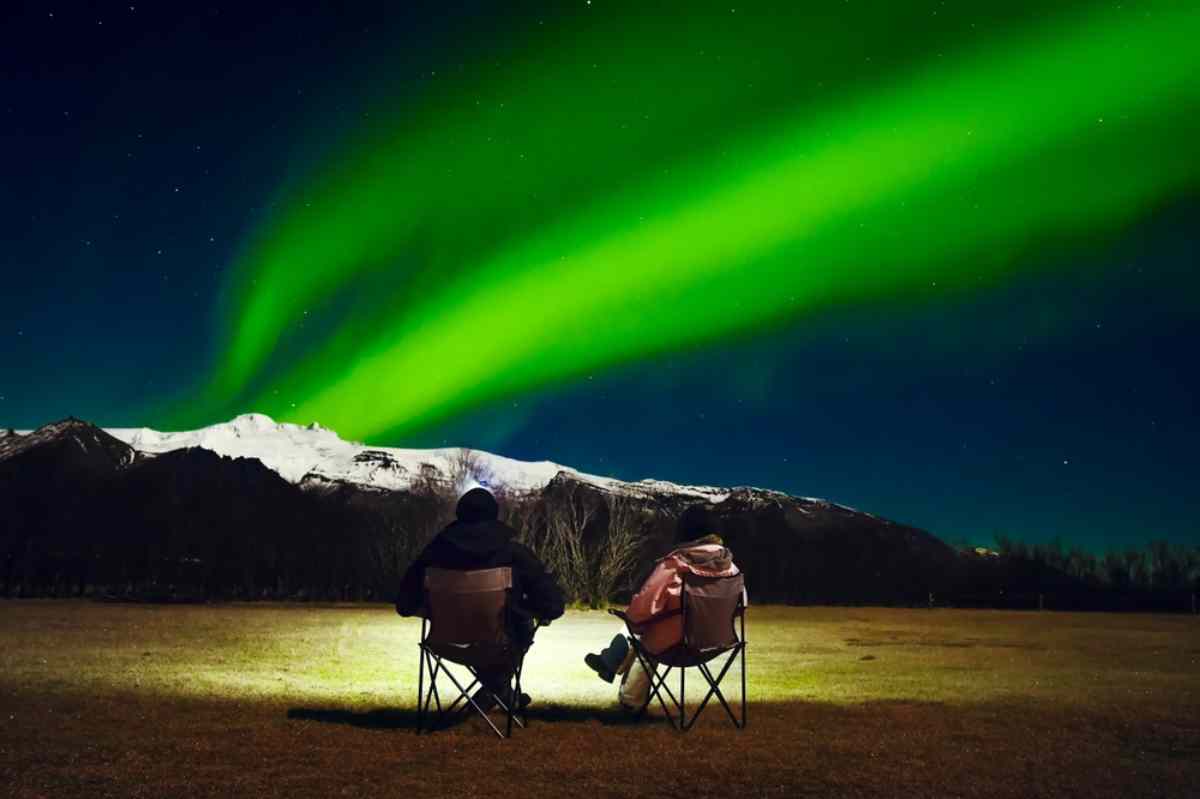 Dos personas sentadas en sillas de camping, contemplando un vibrante espectáculo de auroras boreales de color verde sobre un fondo de montañas cubiertas de nieve bajo una noche clara y estrellada.