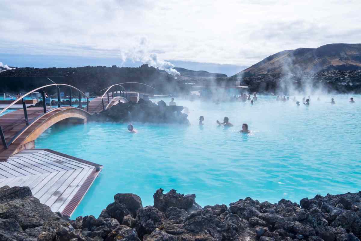 Gente relajándose en las cálidas aguas de color azul lechoso de una fuente termal geotermal, rodeada de rocas de lava negra y puentes de madera, con vapor elevándose sobre un fondo de escarpadas colinas volcánicas.