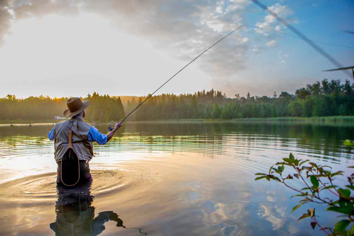 Un pescador de pie hasta las rodillas en un lago sereno, lanzando su caña al amanecer, con el cálido resplandor de la luz de la mañana reflejándose en el agua y rodeado de exuberante vegetación.