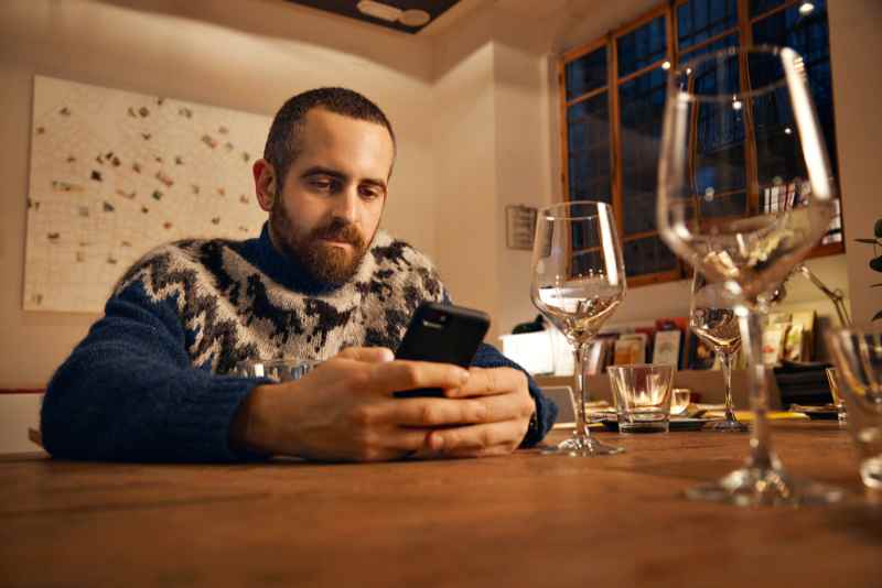 Hombre con su&eacute;ter tradicional island&eacute;s sentado en una mesa en un restaurante y usando su tel&eacute;fono inteligente, rodeado de copas de vino.