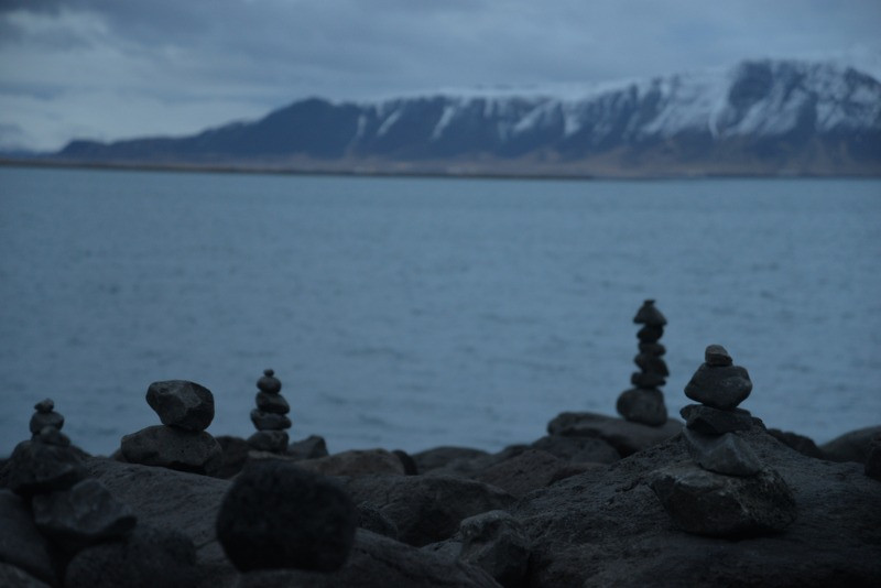 Pequeños cairns de piedra apilados a lo largo de una costa rocosa con un mar en calma y montañas nevadas al fondo bajo un cielo nublado en Islandia.