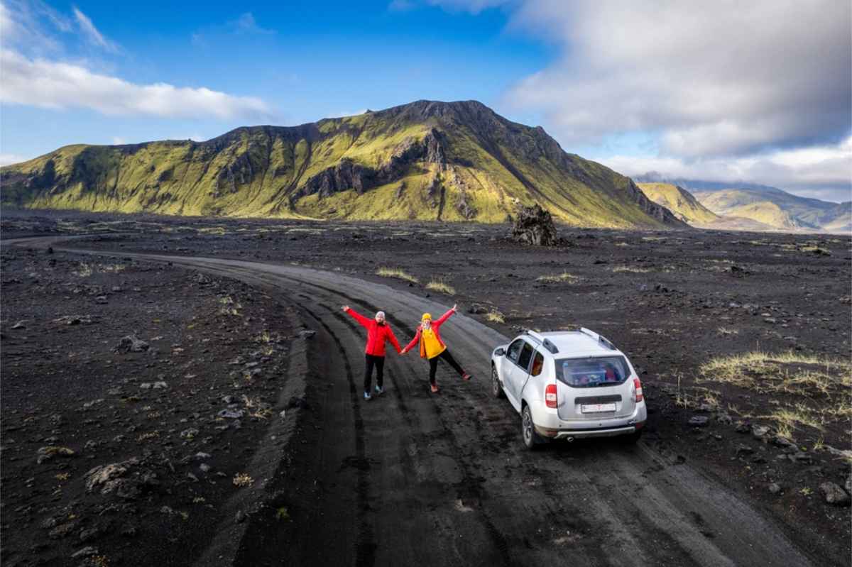 Consejos para reservar un coche de alquiler en Islandia Dos turistas saludando al dron con una vista impresionante del paisaje islandés y su coche de alquiler