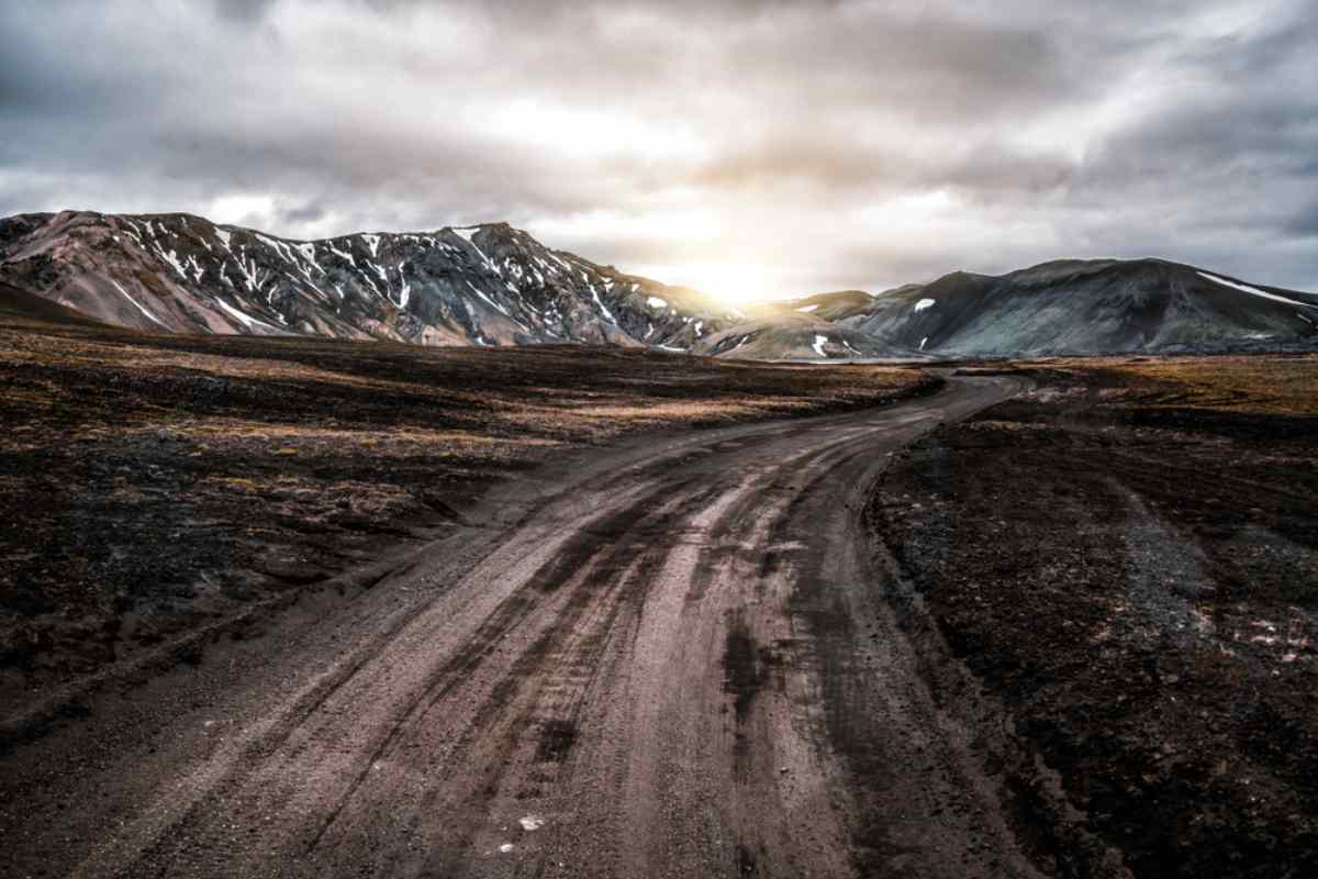 Carretera de grava que serpentea entre montañas islandesas con parches de nieve bajo un cielo invernal dramático.