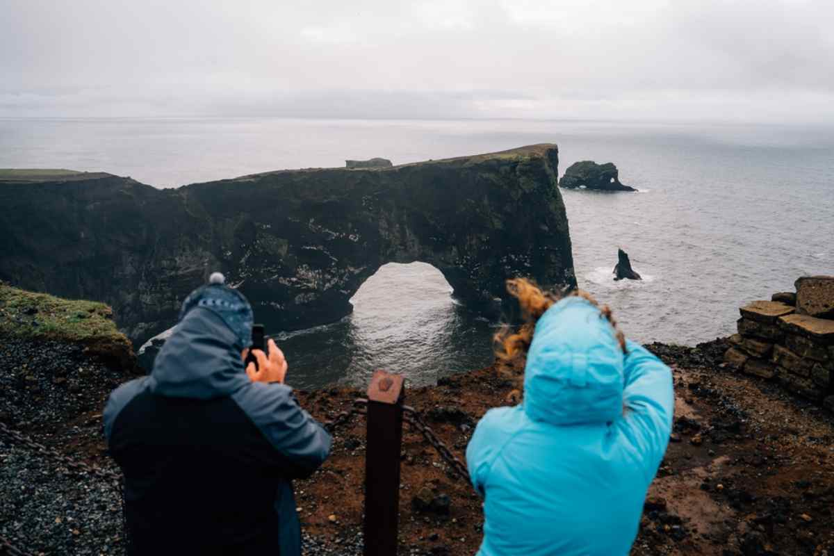 Dos viajeros con chaquetas impermeables fotografiando un impresionante arco marino y acantilados de Islandia en una costa ventosa.