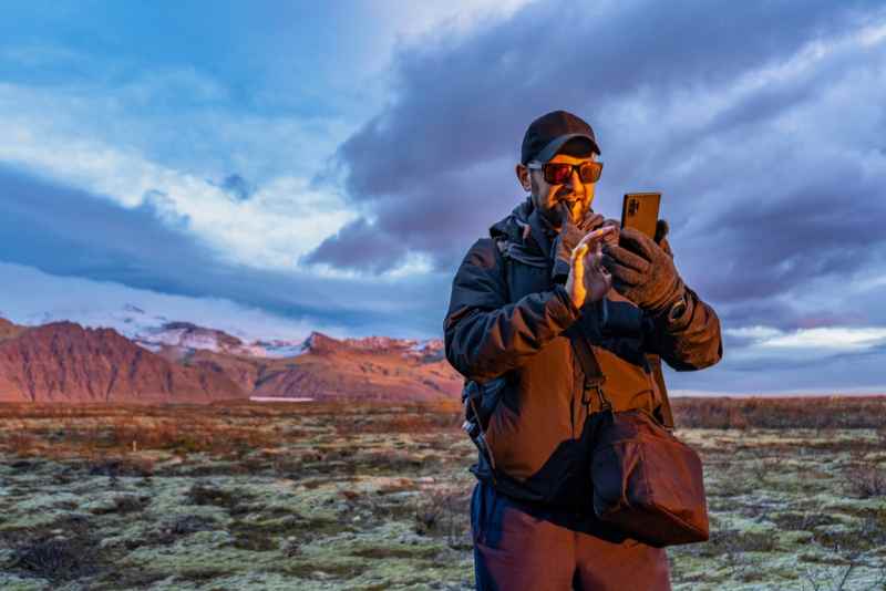 Homme en v&ecirc;tements d'ext&eacute;rieur utilisant son smartphone devant un paysage pittoresque en Islande au coucher du soleil.