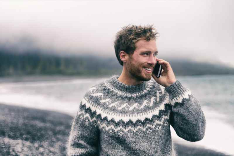 Homme souriant en pull en laine islandais traditionnel parlant au t&eacute;l&eacute;phone sur une plage brumeuse en Islande.