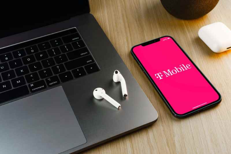Smartphone avec le logo T-Mobile sur l'&eacute;cran &agrave; c&ocirc;t&eacute; d'un ordinateur portable et d'&eacute;couteurs sans fil sur une table en bois.
