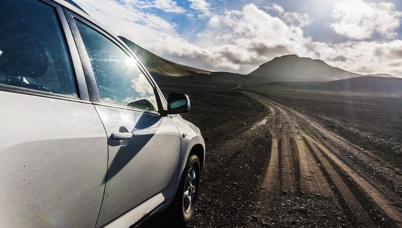 Un SUV sur une route volcanique en Islande – Une aventure épique hors route Un SUV blanc roule sur un chemin de terre à travers le paysage volcanique de l'Islande, avec une montagne au loin sous un ciel ensoleillé rempli de nuages.