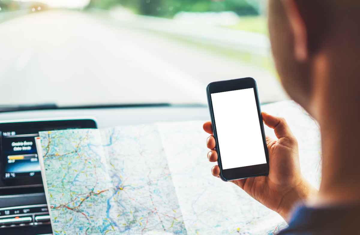Voyageur Utilisant Smartphone et Carte en Papier pour la Navigation - Préparation d'un Road Trip Une personne en voiture tenant un smartphone avec un écran vide tout en regardant une carte papier étalée sur ses genoux, utilisant les deux outils pour la navigation.
