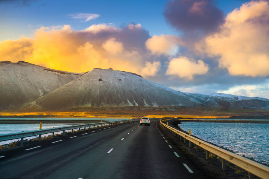 Voiture sur une route côtière en Islande, entre eau et montagnes, ciel doré au coucher du soleil.