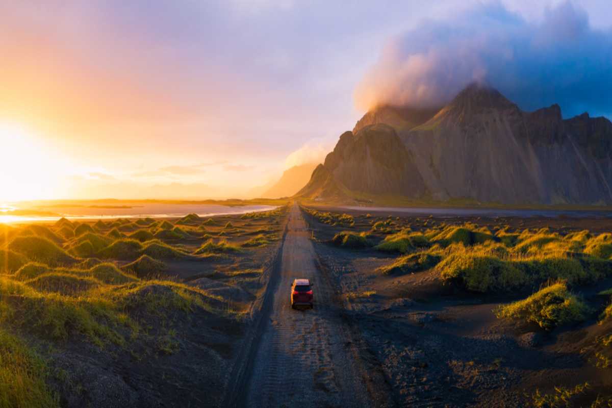 Guidare sulla Costa Sud dell'Islanda con un'auto a noleggio Auto rossa che guida su una strada di sabbia nera lungo la Costa Sud dell’Islanda con montagne spettacolari e luce del tramonto